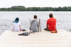 Kaksi nuorta ja aikuinen istuu laiturilla järven luona selin kameraan.